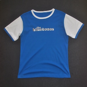 Puede incluir: Camiseta azul con mangas blancas y cuello blanco. El texto "the bluetones" está impreso en plata en el pecho. La camiseta está hecha de un material suave y ligero.