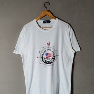 May include: White t-shirt with a circular graphic featuring a compass with the text "Ocean Challenge Polo Sport Ralph Lauren Est. 1967" and a small American flag.