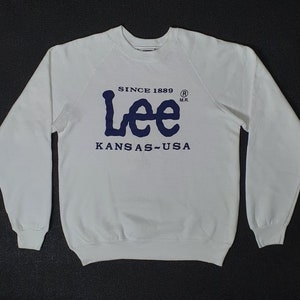 May include: A white sweatshirt with long sleeves. The navy blue text on the front reads "Lee" with "Since 1889" above and "Kansas - USA" below. The sweatshirt is made of a soft material.