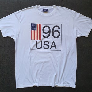 May include: White t-shirt with a graphic print of the American flag, the number 96, and the word "USA".