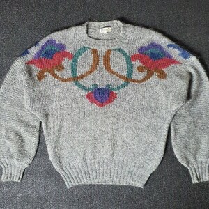 May include: A gray knitted sweater with a floral design in shades of red, blue, green, and brown. The sweater has long sleeves and a crew neck.