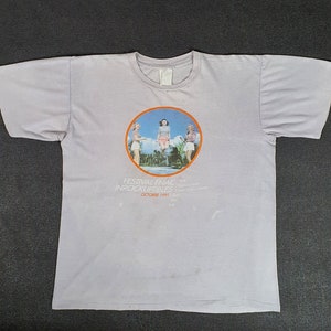 May include: A light purple t-shirt with a circular graphic of three women in swimsuits jumping in the air. The graphic is surrounded by text that reads "Festival Final - Inrockuptibles - October 1981".