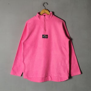 Puede incluir: Un jersey de forro polar rosa con cierre de cremallera de un cuarto y un pequeño logotipo colorido en la parte delantera.