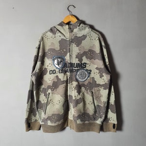 Könnte beinhalten: Ein Kapuzenpullover in Tarnmuster mit Reißverschluss. Der Pullover hat zwei Aufnäher auf der Vorderseite, einen mit einem Vogel und dem Text "Since 1974" und den anderen mit dem Text "ADIUMS CO. CHAMPIONS" und einem Logo.