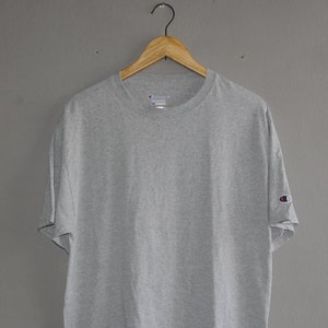 Puede incluir: Una camiseta gris de manga corta con un logotipo de Champion en la manga izquierda.