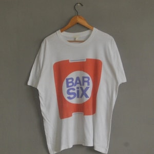 May include: White t-shirt with a red and purple logo that says "Bar Six" on it.