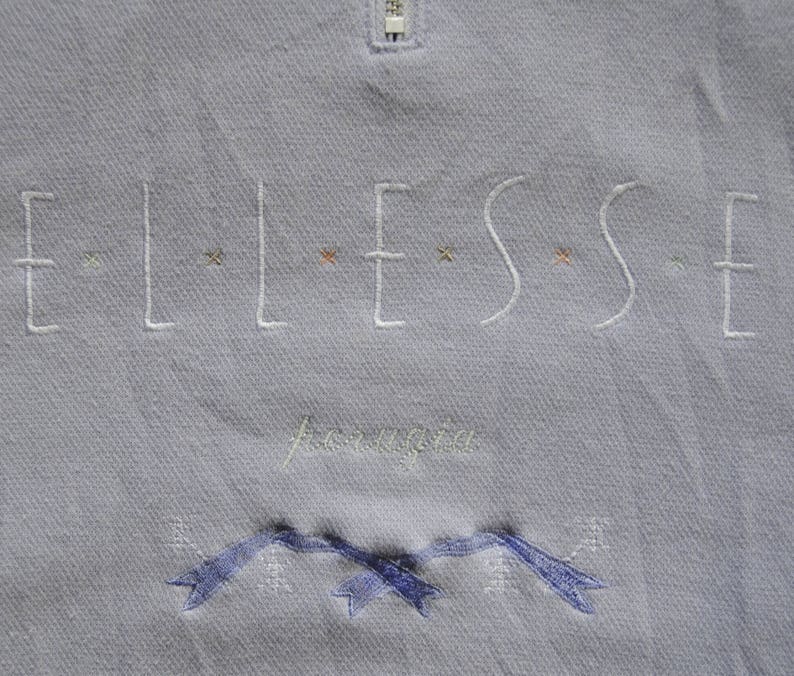 May include: A light blue sweatshirt with the word "ELLESSE" embroidered in white with small colored accents. The word "Aeronautica" is embroidered in white below the logo. The sweatshirt has a zipper at the top.