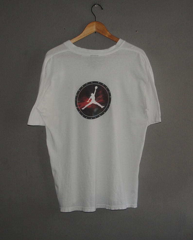 K&ouml;nnte beinhalten: Wei&szlig;es T-Shirt mit einem schwarzen und roten kreisf&ouml;rmigen Grafik eines Basketballspielers, der einen Dunk macht. Die Grafik enth&auml;lt den Text "Air Jordan" und eine Silhouette des Jumpman-Logos von Michael Jordan.