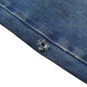 May include: Close-up of blue denim fabric with a distressed hole. The denim shows signs of wear and tear, with frayed edges around the hole. The fabric is a medium blue color.