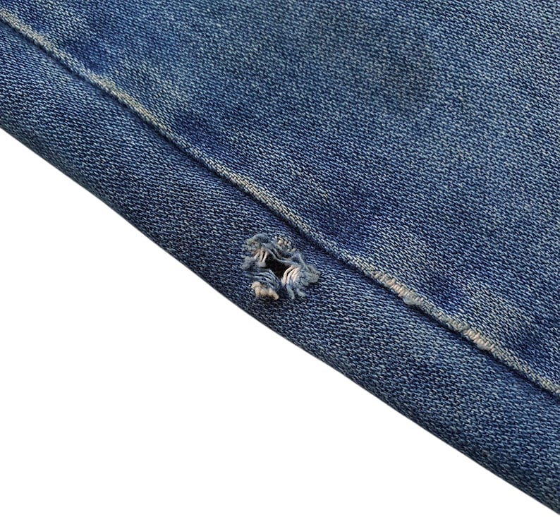 May include: Close-up of blue denim fabric with a distressed hole. The denim shows signs of wear and tear, with frayed edges around the hole. The fabric is a medium blue color.
