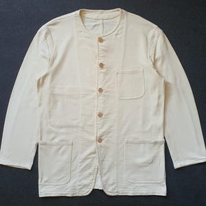 May include: A long-sleeved, off-white jacket featuring a collarless round neckline and a front button closure with five light-colored buttons. The garment includes one chest pocket on the left side and two larger patch pockets at the bottom hem.