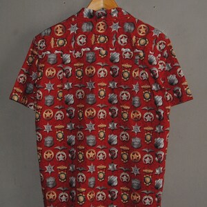 Pode incluir: Camisa vermelha de manga curta com um padr&atilde;o repetido de distintivos de xerife, estrelas e outros emblemas policiais. A camisa est&aacute; pendurada em um cabide de madeira contra um fundo cinza.