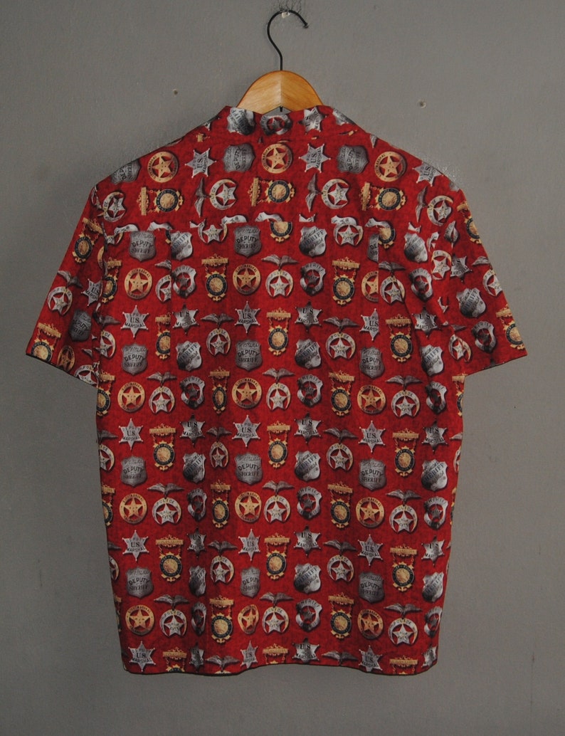 Pode incluir: Camisa vermelha de manga curta com um padr&atilde;o repetido de distintivos de xerife, estrelas e outros emblemas policiais. A camisa est&aacute; pendurada em um cabide de madeira contra um fundo cinza.