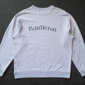 May include: Light gray sweatshirt with the word "Pendleton" arched across the back in dark gray lettering. The long-sleeved crew neck shirt has gray ribbed cuffs and hem. The fabric appears to be a soft, heathered material.