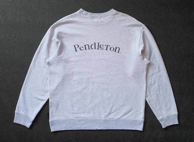 May include: Light gray sweatshirt with the word "Pendleton" arched across the back in dark gray lettering. The long-sleeved crew neck shirt has gray ribbed cuffs and hem. The fabric appears to be a soft, heathered material.