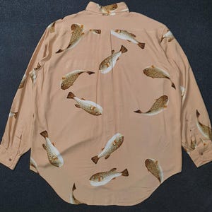 K&ouml;nnte beinhalten: Hellpfirsichfarbenes Langarmhemd mit einem sich wiederholenden Muster aus braunen und wei&szlig;en Fischen. Das Hemd hat einen klassischen Kragen und gekn&ouml;pfte Manschetten. Das Fischmuster ist &uuml;ber den Stoff verteilt.