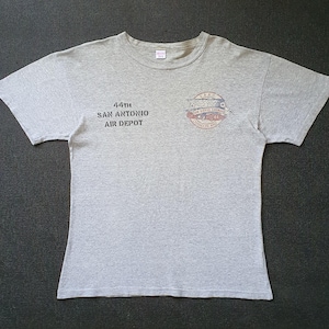 Può includere: Una t-shirt grigio melange a maniche corte con la scritta 44TH SAN ANTONIO AIR DEPOT stampata in nero sul petto sinistro. A destra del testo si trova un emblema grafico circolare con dettagli rossi e blu e altro testo piccolo.