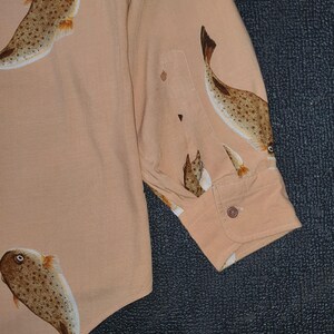 K&ouml;nnte beinhalten: Hellbeige Hemd mit Kn&ouml;pfen und einem sich wiederholenden Muster aus braunen und wei&szlig;en Fischen. Das Hemd hat eine Tasche und eine Manschette mit Knopf. Das Fischdesign ist detailliert mit Punkten und Flossen.