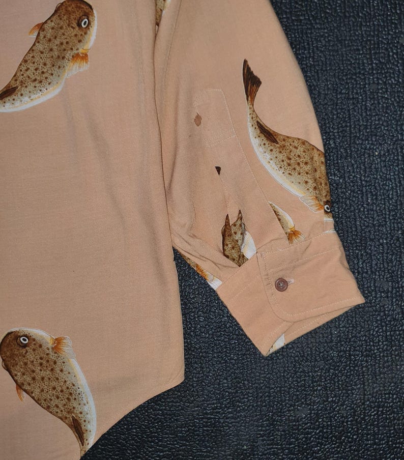 K&ouml;nnte beinhalten: Hellbeige Hemd mit Kn&ouml;pfen und einem sich wiederholenden Muster aus braunen und wei&szlig;en Fischen. Das Hemd hat eine Tasche und eine Manschette mit Knopf. Das Fischdesign ist detailliert mit Punkten und Flossen.