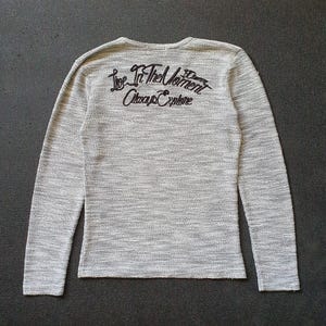 May include: A light gray long-sleeve shirt with a textured knit pattern. The back of the shirt features black embroidered text that reads "Live In The Moment Always Explore". The shirt has a crew neck.