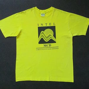 Può includere: Una t-shirt a maniche corte giallo neon con una grafica quadrata nera sul petto. La grafica mostra il testo INTEL MCD sopra un design stilizzato di montagna e sole, con la frase A GREAT PLACE TO MAKE MEMORIES stampata sotto.