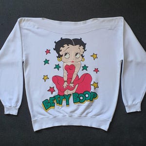 Op de afbeelding: Witte sweatshirt met een Betty Boop-afbeelding. Het stripfiguur draagt een rode jurk, omringd door sterren, met de tekst "Betty Boop" in groen en geel. De top met lange mouwen heeft een ronde hals.