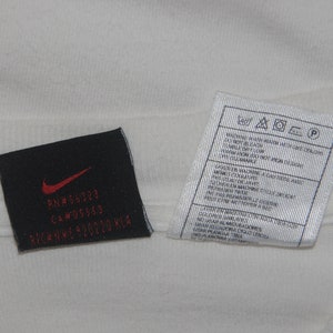 K&ouml;nnte beinhalten: Ein wei&szlig;es T-Shirt mit einem schwarzen Pflegeetikett und einem schwarzen Etikett mit dem Text "RN# 56323 CA# 05553  NIKE HWY 920220 KLA"
