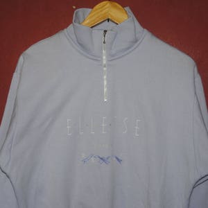 May include: A light blue quarter-zip sweatshirt with the word "ELLESSE" embroidered on the front. The sweatshirt has a small mountain graphic below the word.