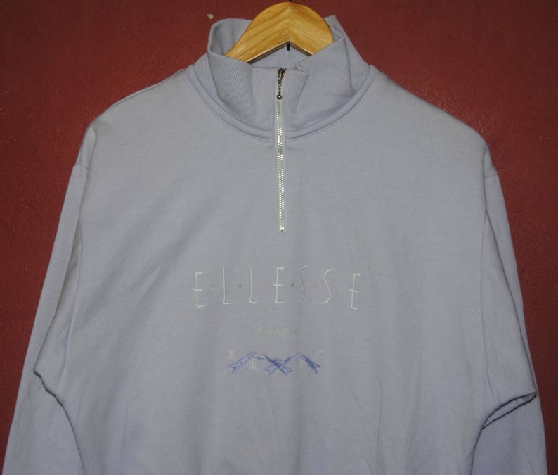 May include: A light blue quarter-zip sweatshirt with the word "ELLESSE" embroidered on the front. The sweatshirt has a small mountain graphic below the word.