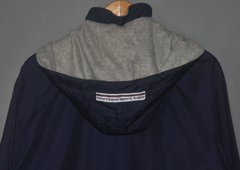 Puede incluir: Una chaqueta azul marino con capucha y forro de polar gris. La chaqueta tiene una etiqueta blanca y roja con el texto "Courreges Sport Futur".