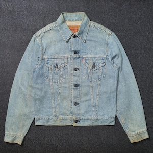 Op de afbeelding: Lichtblauwe spijkerjas met knoopsluitingen, twee borstzakken en een klassieke kraag. De jas heeft een verweerde uitstraling, wat duidt op slijtage. Een klein rood label is zichtbaar op de borstzak.