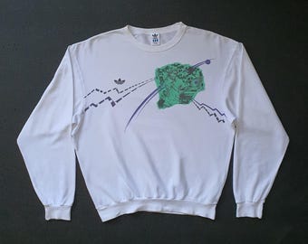 May include: White long-sleeve sweatshirt with a graphic design. The design features a green and blue image with purple lines and mountain-like shapes. The Adidas logo is visible on the upper left chest and on the tag.