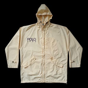 May include: A light yellow hooded jacket with the text "POLO 1992" in navy blue. The jacket has a full zipper, two front pockets, and buttoned cuffs. The jacket is made of a lightweight material and is suitable for outerwear.