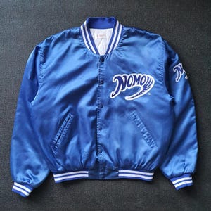 Puede incluir: Chaqueta bomber de satén azul real con ribete a rayas blancas y azules en el cuello, los puños y el dobladillo. La chaqueta presenta la palabra "NOMO" en escritura blanca en el pecho y un logotipo a juego en la manga. Se ven dos bolsillos delanteros.
