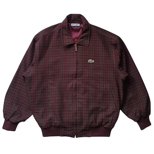 May include: A burgundy and black plaid zip-up jacket featuring a collared neckline, long sleeves with ribbed cuffs, a ribbed hem, and a small green crocodile logo embroidered on the left chest. The interior is lined with a solid burgundy fabric.