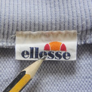 May include: A white label with the word "ellesse" in blue lettering and a red and orange logo of a half circle with a red stripe across the top. The label is attached to a light blue ribbed fabric.