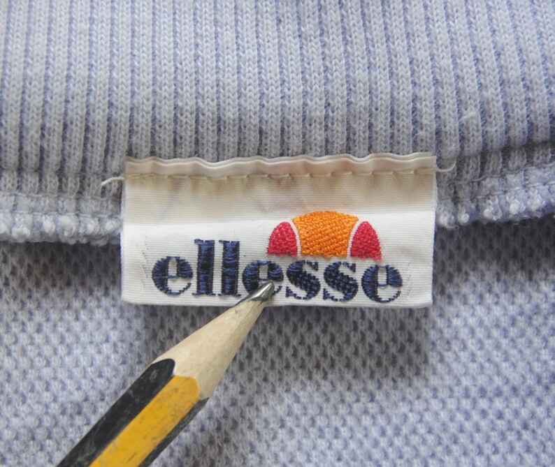 May include: A white label with the word "ellesse" in blue lettering and a red and orange logo of a half circle with a red stripe across the top. The label is attached to a light blue ribbed fabric.