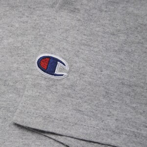 Op de afbeelding: Een grijs T-shirt met een geborduurd Champion-logo in blauw en rood op de linkerborst.