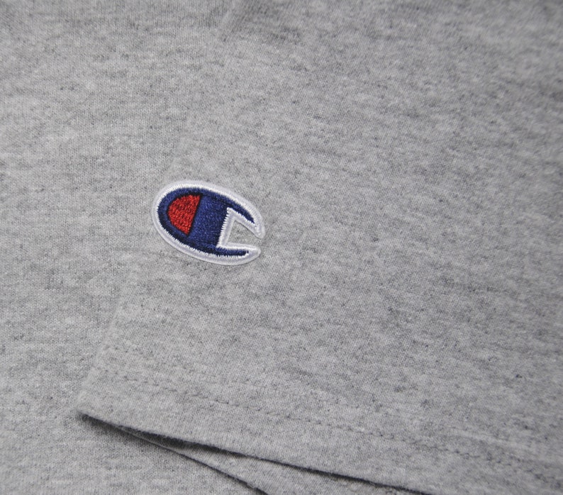 Op de afbeelding: Een grijs T-shirt met een geborduurd Champion-logo in blauw en rood op de linkerborst.