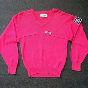 Könnte beinhalten: Ein leuchtend pinkfarbener V-Ausschnitt-Pullover mit langen Ärmeln. Der Pullover hat Rippendetails an Brust und Bündchen sowie kleine weiße rechteckige Aufnäher mit Text. Ein Aufnäher am Oberarm trägt die Aufschrift "RACING SPORTS DIVISION".