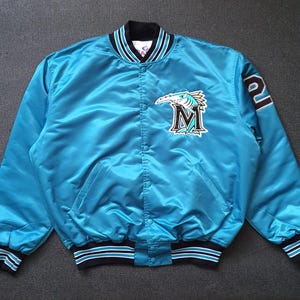 May include: A bright teal satin bomber jacket featuring a snap-button front closure, long sleeves, and ribbed knit cuffs and hem with black and white stripes. The chest displays a black and white marlin fish logo with the letter M.
