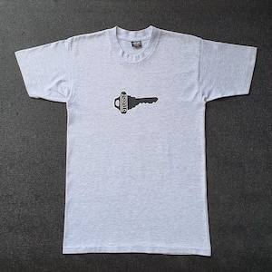 Puede incluir: Camiseta gris claro con un diseño de llave negra. El gráfico de la llave tiene la palabra "Stussy" impresa en la cabeza de la llave. La camiseta está hecha de un material suave y tiene mangas cortas.