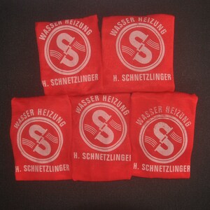 Op de afbeelding: Vijf rode stoffen zakjes met witte tekst en een wit logo van een pijp met water dat erdoorheen stroomt. De tekst op de zakjes luidt "Wasser Heizung" en "H. Schnetzlinger".