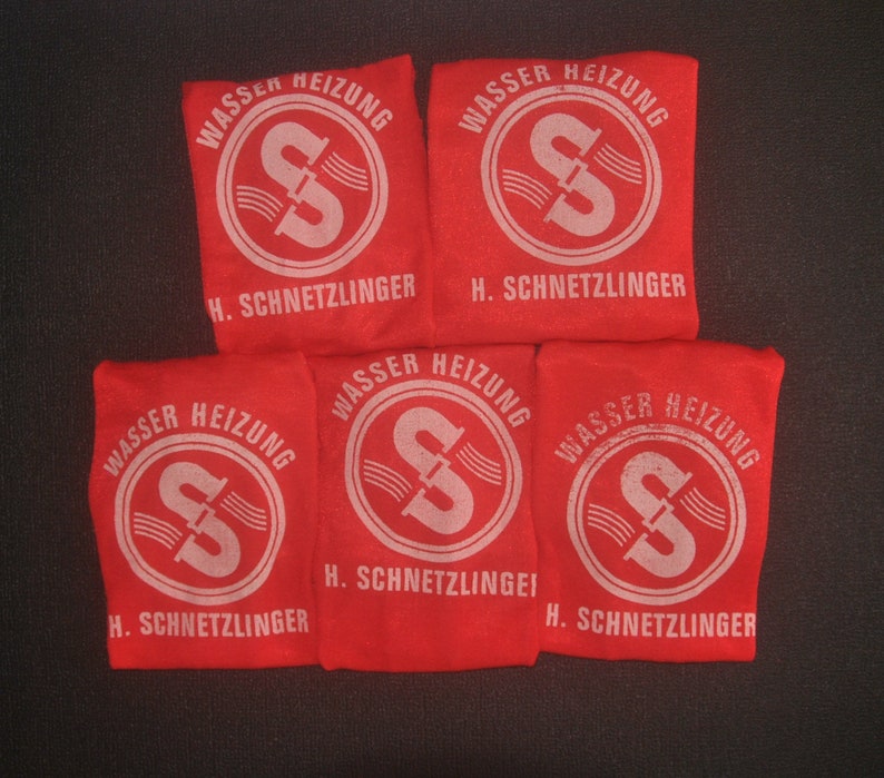Op de afbeelding: Vijf rode stoffen zakjes met witte tekst en een wit logo van een pijp met water dat erdoorheen stroomt. De tekst op de zakjes luidt "Wasser Heizung" en "H. Schnetzlinger".
