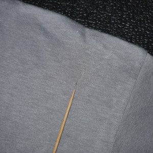 May include: Close-up of a gray heather t-shirt with a small hole near the neckline. The fabric is textured and has a slight sheen.