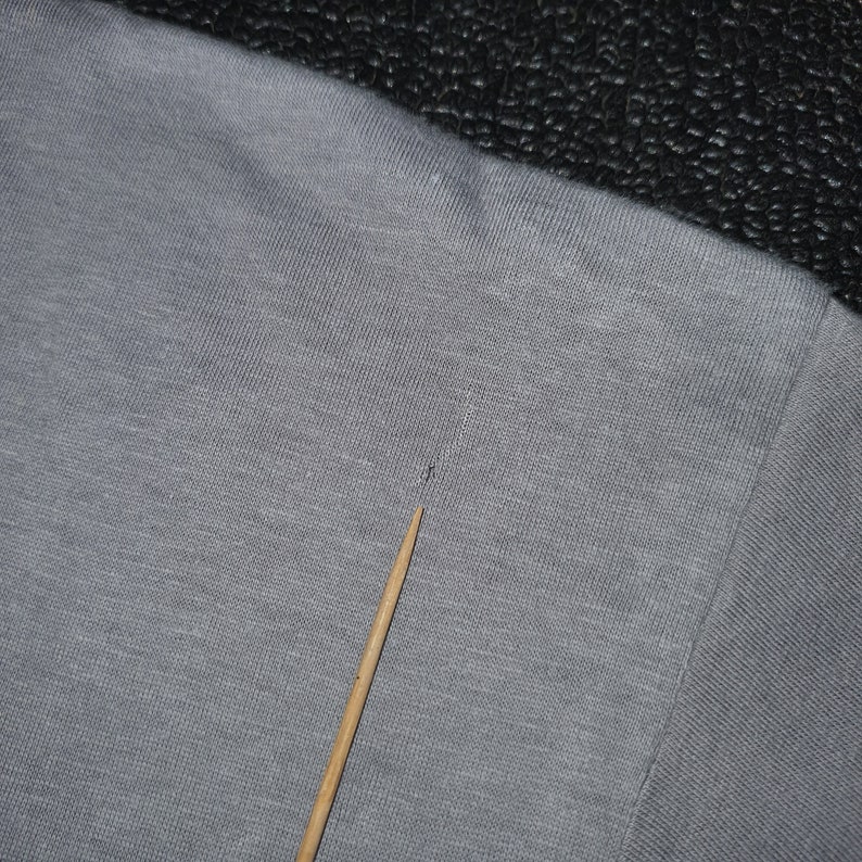 May include: Close-up of a gray heather t-shirt with a small hole near the neckline. The fabric is textured and has a slight sheen.