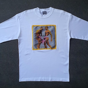May include: White t-shirt with a square graphic depicting two nude male figures. The graphic is framed in yellow and features the brand name "Versace". The shirt has short sleeves and a crew neck.