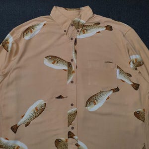 K&ouml;nnte beinhalten: Hellbeige Hemd mit Kn&ouml;pfen und einem sich wiederholenden Muster von Kugelfischen. Das lang&auml;rmlige Hemd hat einen Button-Down-Kragen und eine Brusttasche. Die Kugelfische sind braun mit wei&szlig;en B&auml;uchen.