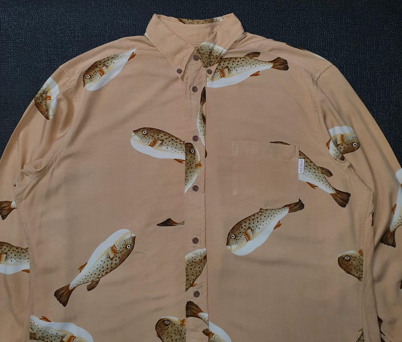 K&ouml;nnte beinhalten: Hellbeige Hemd mit Kn&ouml;pfen und einem sich wiederholenden Muster von Kugelfischen. Das lang&auml;rmlige Hemd hat einen Button-Down-Kragen und eine Brusttasche. Die Kugelfische sind braun mit wei&szlig;en B&auml;uchen.
