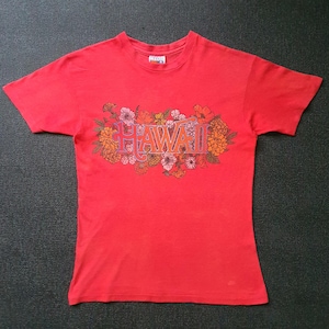 May include: A red t-shirt featuring the word "HAWAII" in a floral design. The text is in shades of orange and pink, surrounded by colorful flowers. The shirt has short sleeves and a crew neck.